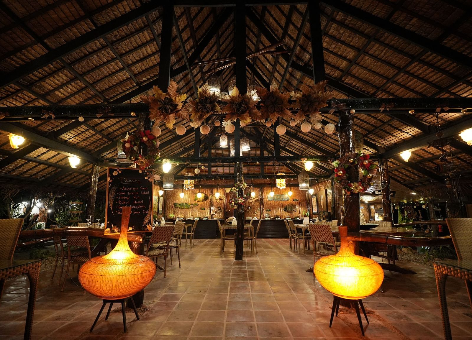 Interior of Flower Island Resort restaurant with wooden furniture and yellow pendant lights under a thatched roof.