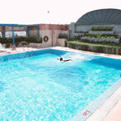 Rooftop swimming pool at The Bristol Hotel, Gurgaon with a person swimming, tiered planters in the background, and a pergola with lounge chairs beside the pool.