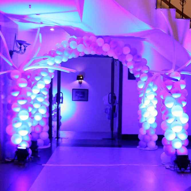 Entrance at The Bristol Hotel, Gurgaon decorated with a white balloon arch styled as palm trees, lit with purple and blue uplighting, leading to a doorway.