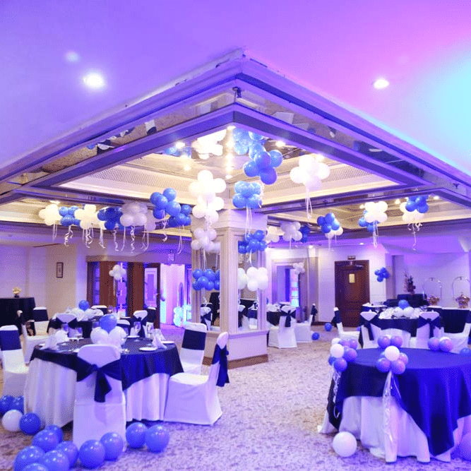 Banquet hall at The Bristol Hotel, Gurgaon with a mirrored ceiling reflecting balloons, round tables in navy and white linen, and buffet stations beyond.