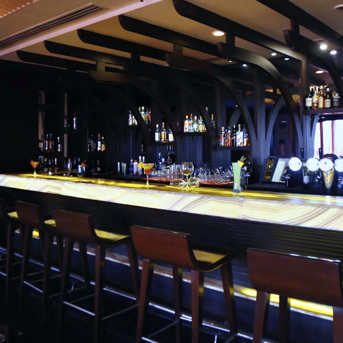 Bar at The Bristol Hotel, Gurgaon with a backlit onyx-style counter, wooden arched ceiling beams, rows of bottles on shelves, and leather bar stools.