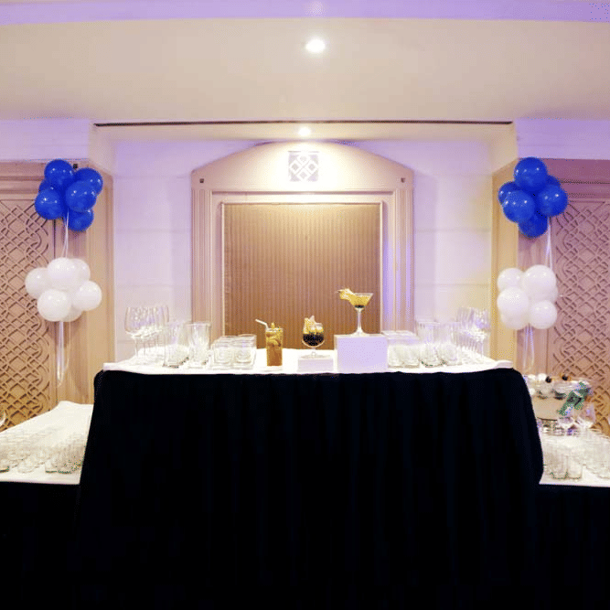Bar setup at The Bristol Hotel, Gurgaon with black-draped tables lined with glassware, carved wooden wall panels, and balloon bunches on either side.