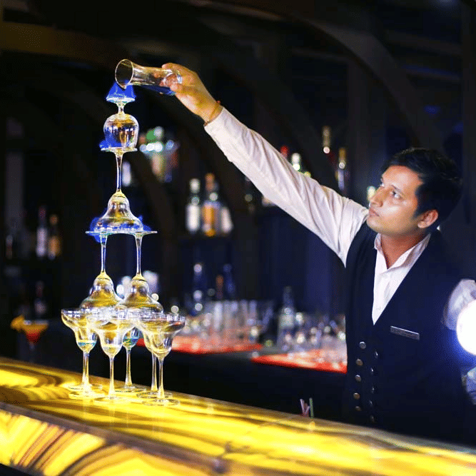 Bartender at The Bristol Hotel, Gurgaon pouring blue liqueur down a tiered champagne glass tower on a backlit bar counter lined with bottles.