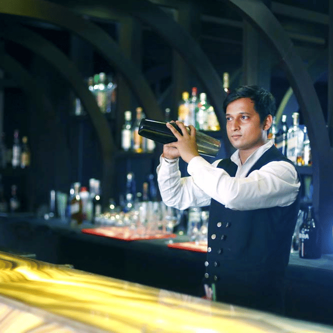 A bartender at The Bristol Hotel, Gurgaon using a cocktail shaker at the illuminated bar counter, with backlit shelves of bottles under arched wooden beams.