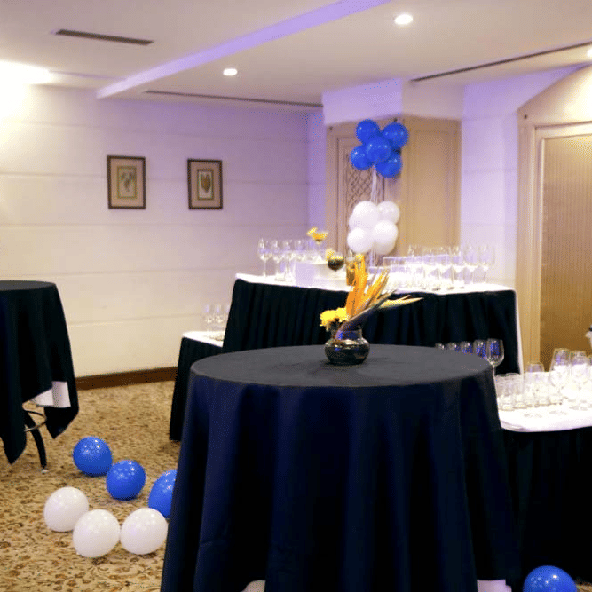 Birthday celebration setup at The Bristol Hotel, Gurgaon with a tiered cake on a black-draped table, superhero backdrop cutout, balloons under purple lighting.