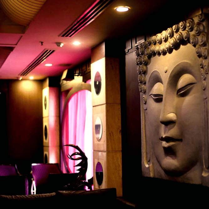 A lounge at The Bristol Hotel, Gurgaon featuring a large carved Buddha wall relief, pink ambient lighting, purple velvet seating, and wooden pillars.