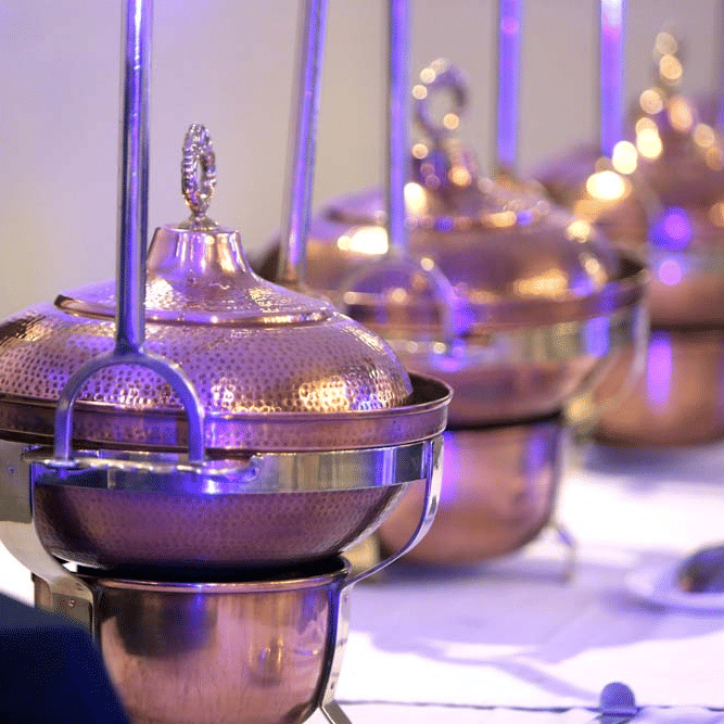 Buffet setup at The Bristol Hotel, Gurgaon with a row of hammered copper chafing dishes with domed lids on a white-clothed table under purple lighting.