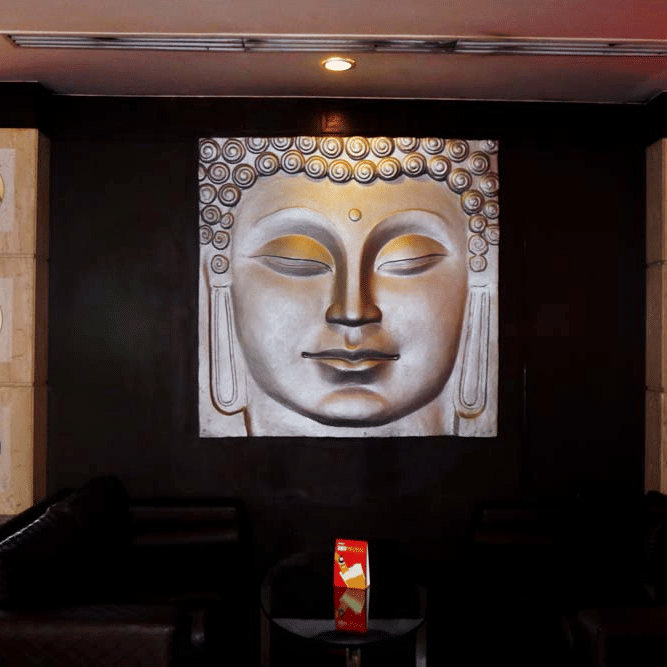 A lounge at The Bristol Hotel, Gurgaon with a golden Buddha wall painting, dark leather sofas, a round table, and stone pillars with circular inlays.
