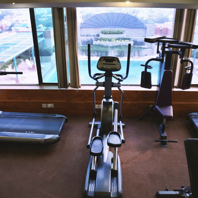 Gym at The Bristol Hotel, Gurgaon with treadmills, an elliptical trainer, and a weight machine in front of floor-to-ceiling windows overlooking the pool.