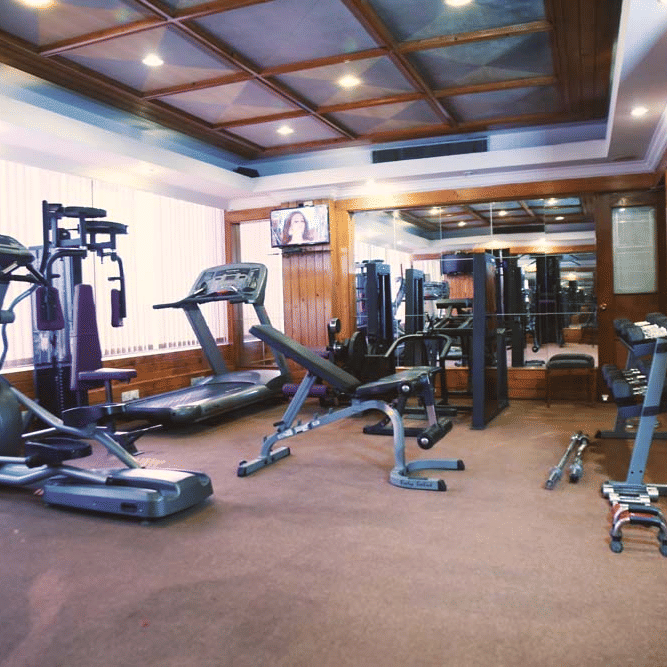 Gym at The Bristol Hotel, Gurgaon with an elliptical trainer, treadmill, weight bench, mirrored wall, wood-panelled ceiling, and a wall-mounted television.