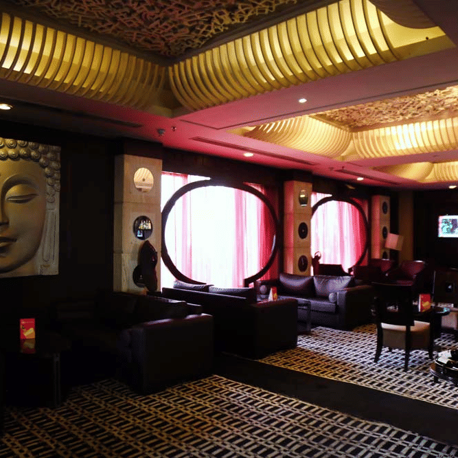A wide view of the lounge at The Bristol Hotel, Gurgaon with a golden Buddha wall painting, a ribbed ceiling design, dark leather sofas, and patterned carpet.