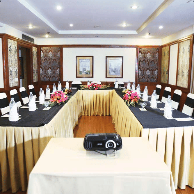 Meeting room at The Bristol Hotel, Gurgaon with a U-shape table in gold and black linen, a projector in the foreground, and 2 framed pictures on the wall.