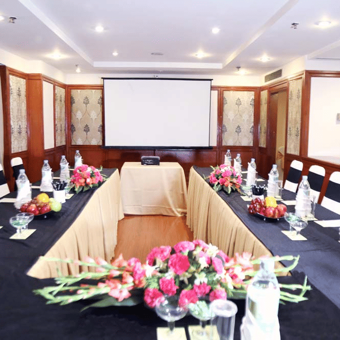 Meeting room at The Bristol Hotel, Gurgaon with tables arranged facing a projector screen, black chair covers, and pink flower arrangements on the tables.
