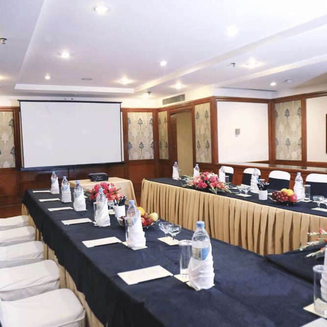 Meeting room at The Bristol Hotel, Gurgaon set in U-shape with navy and gold table linens, white chair covers, a projector screen, and flower arrangements.