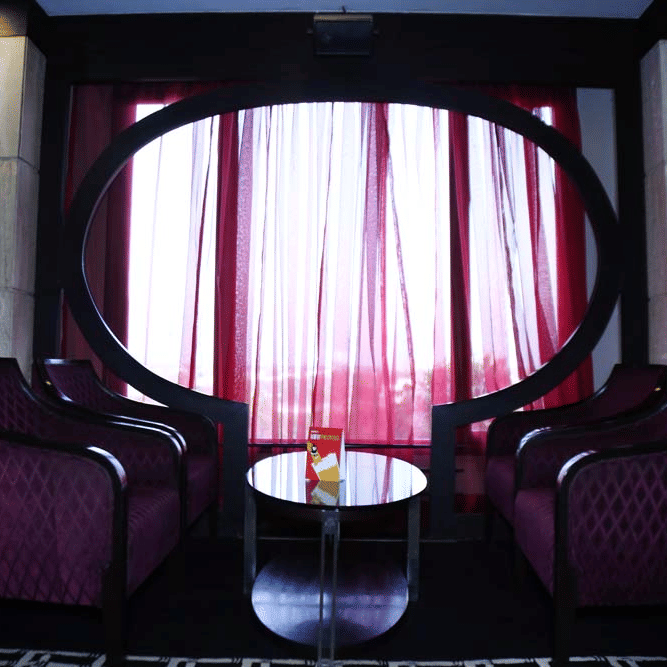 A seating area at The Bristol Hotel, Gurgaon with purple diamond-quilted armchairs, a round table, an oval black frame, and burgundy sheer curtains.