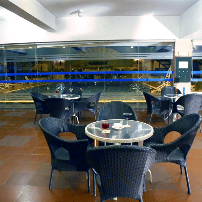 Poolside lounge at The Bristol Hotel, Gurgaon at night with wicker chairs around round glass tables and a view through glass walls to the illuminated pool.