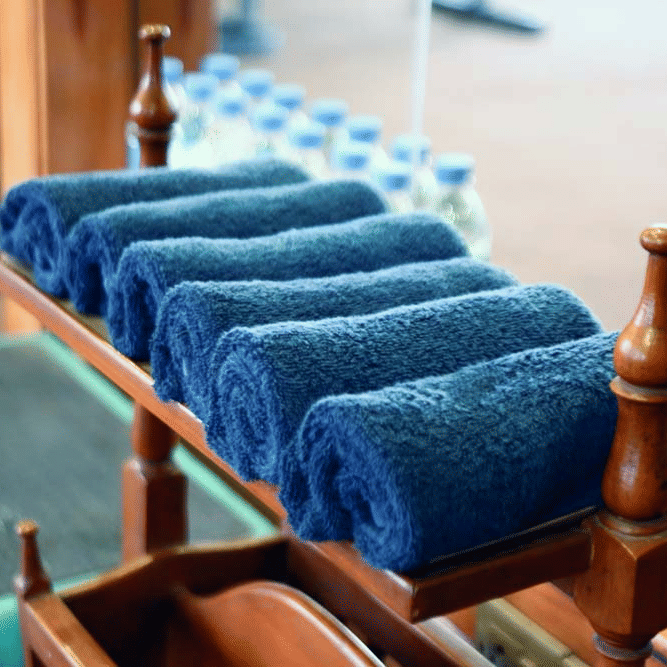Rolled navy blue towels stacked on a wooden poolside trolley at The Bristol Hotel, Gurgaon with small water bottles visible in the background.