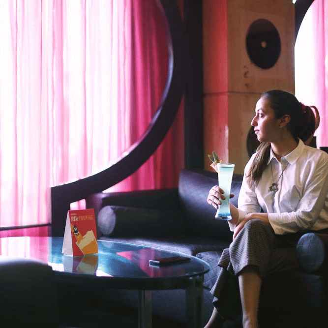 A woman seated on a lounge sofa at The Bristol Hotel, Gurgaon holding a cocktail, with pink curtains and a coffee table with a table tent card.
