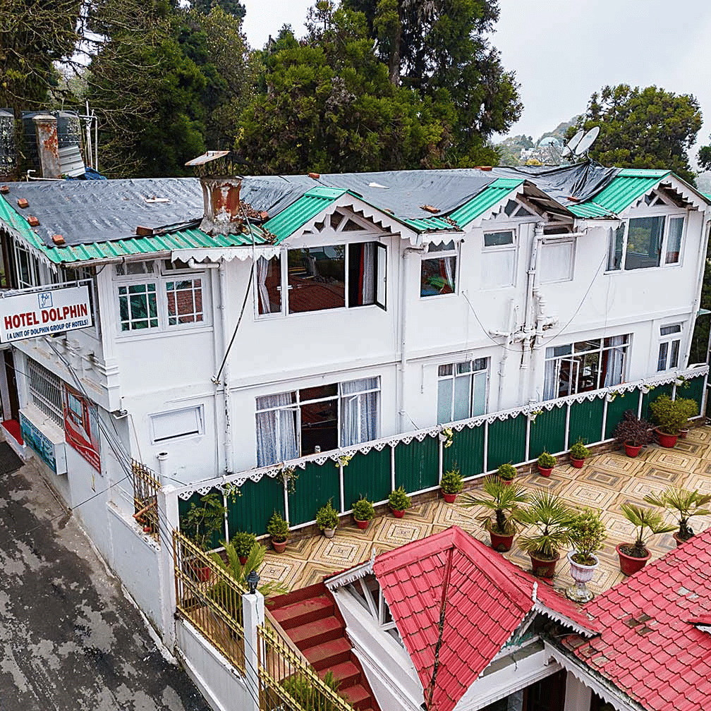 An aerial view of a multi-level white hotel building with green and red roofs nestled among trees on a hillside at Hotel Dolphin Darjeeling.