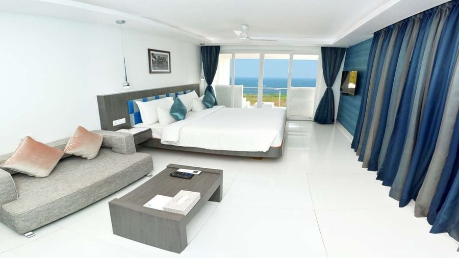 A modern hotel room with a large bed, a grey sofa, and a coffee table, featuring floor-to-ceiling windows with blue curtains offering a view of the ocean | Grande Chalet Sea View | Grand Bay Resort and Spa