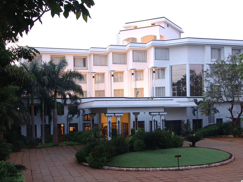 Facade view of Sangam Hotel, Thanjavur, with a pathway, a manicured garden and trees in view - Thanjavur Hotel Contact