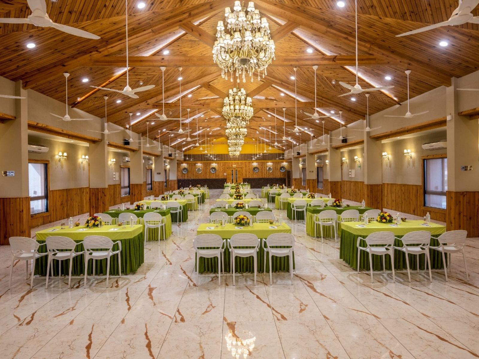 A well-lit banquet hall with multiple tables and chairs set for an event at Angel Lords Eco Inn Amreli.