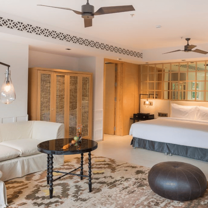 The private pool villa in Goa with a pouffe and a coffee table surrounded by a white couch in front of a plush bed at Azaya Beach Resort Goa