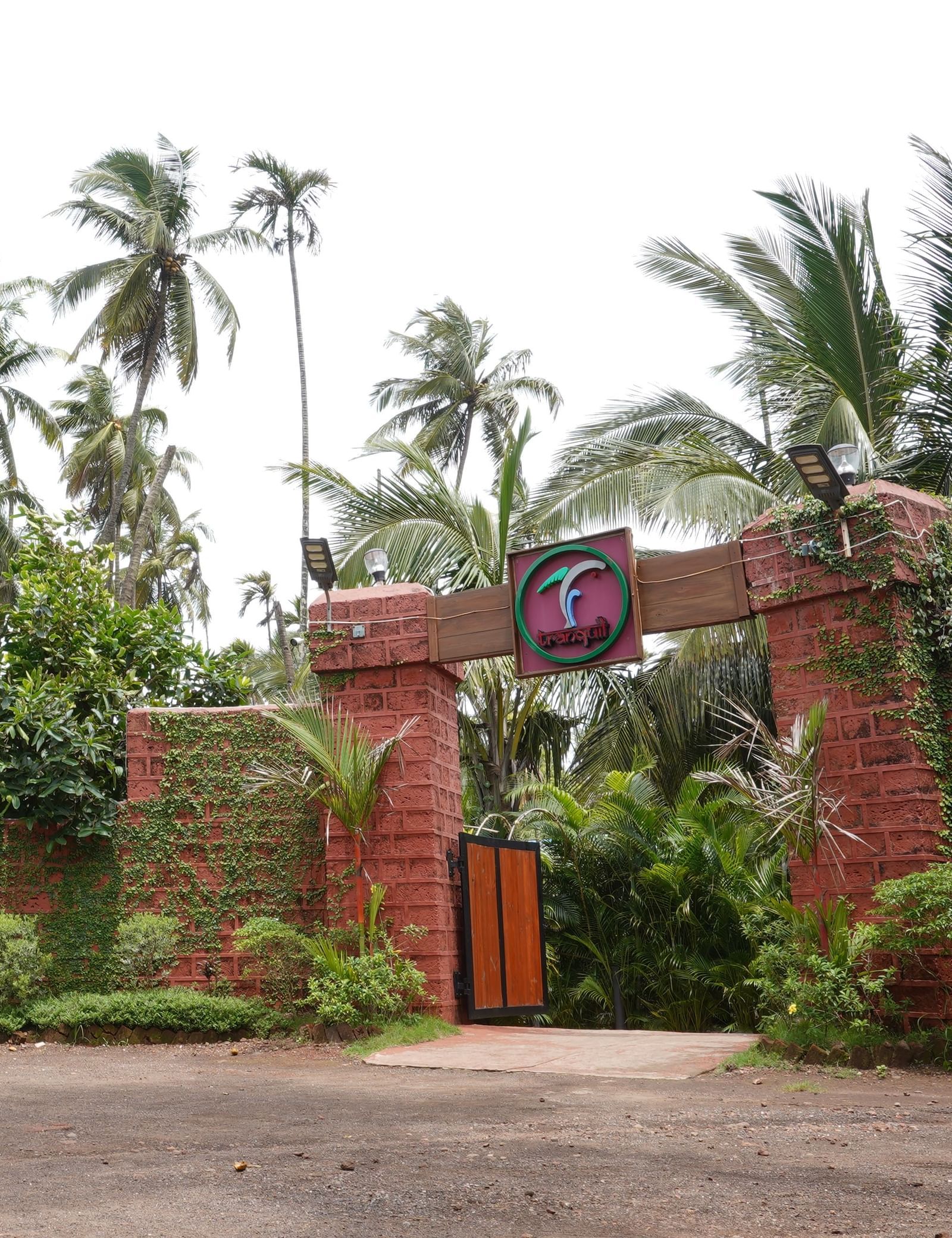 Resort entrance showing a red brick archway, a vibrant sign, and surrounded by coconut trees - Tranquil Beach Resort, Harihareshwar