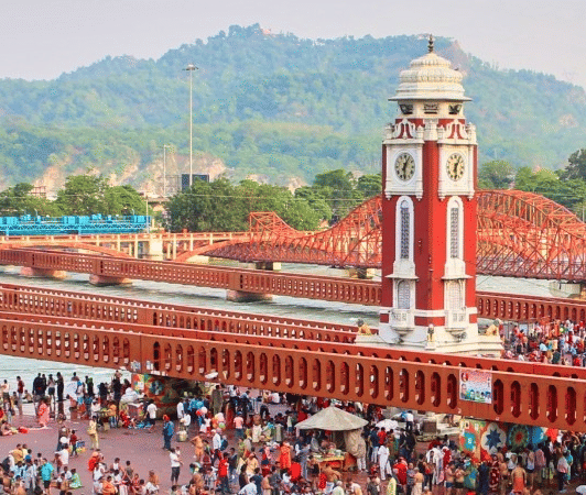 Crowds gather near Har Ki Pauri ghat in Haridwar, with the clock tower and mountains visible in the background