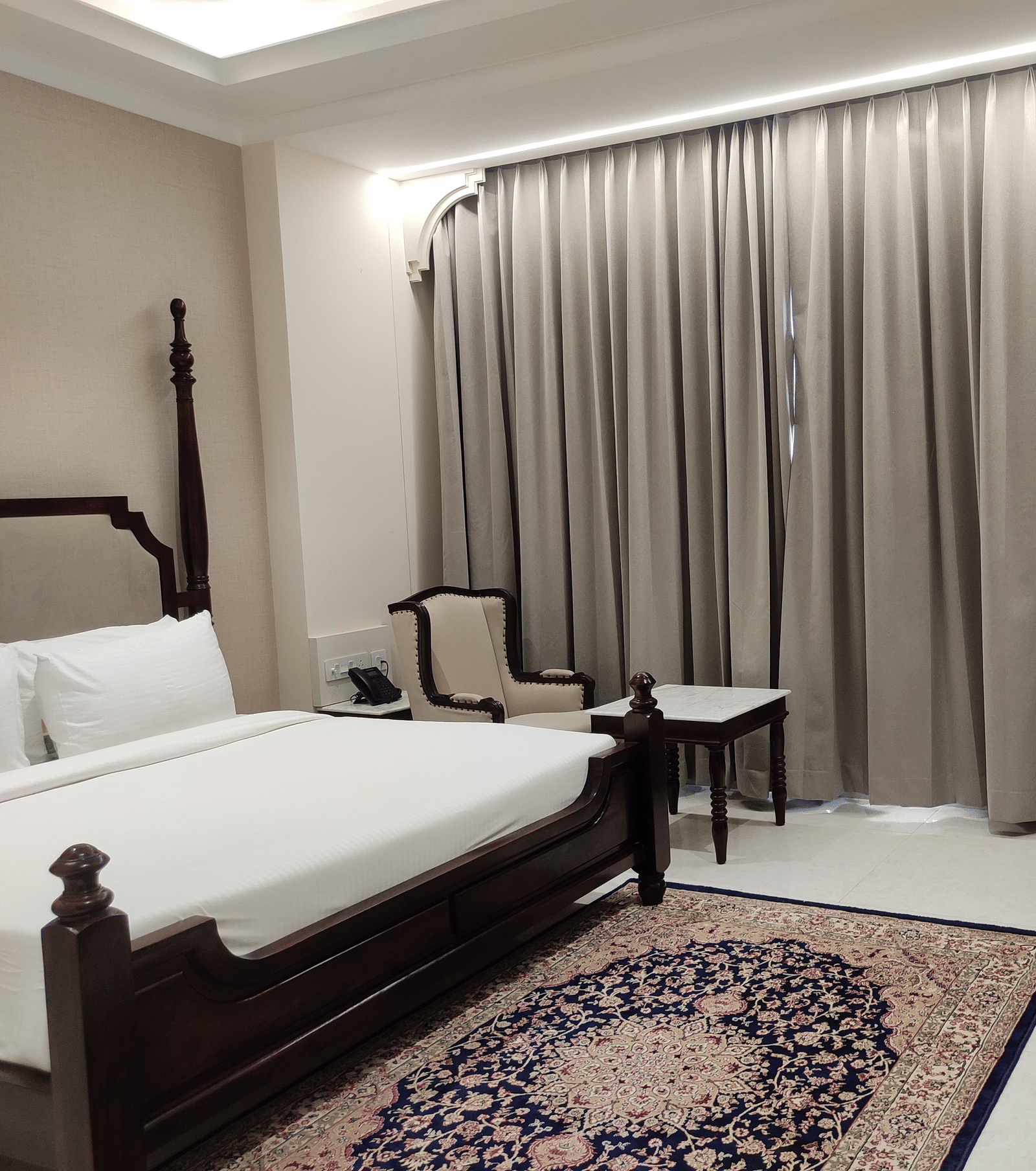 A room interior at Anant Mahal, Jaipur featuring a king size bed, a patterned rug on the floor, a wall mounted television, and long curtains covering a large window behind the seating area.