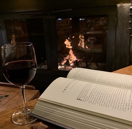 A cozy setting with an open book and a glass of wine placed on a wooden table near a fireplace, creating a warm and relaxing ambiance.