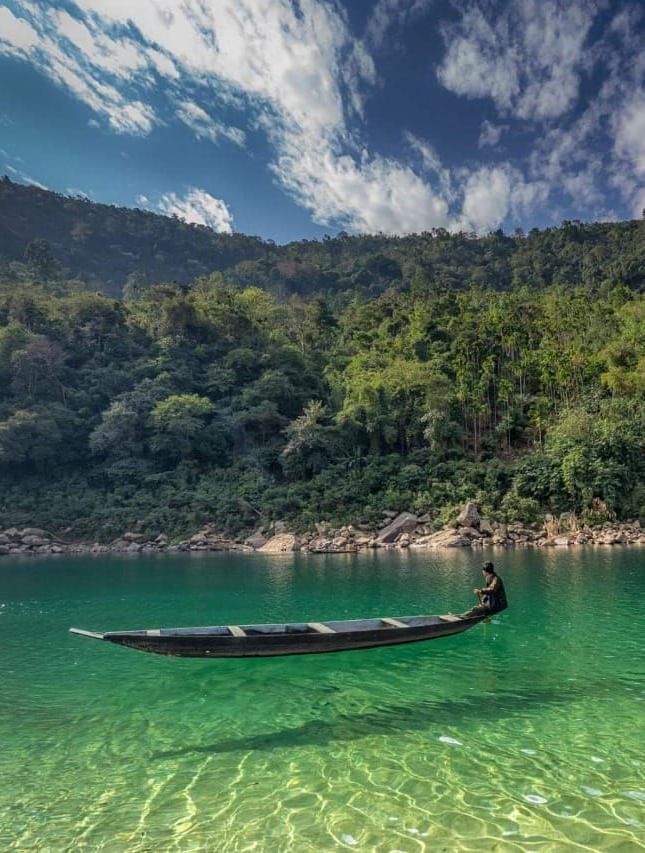 A serene river surrounded by lush forest with a lone wooden boat floating on the water - Polo Hotel Shillong