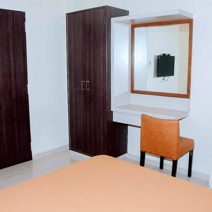 Deluxe room interior featuring a wooden door, dark brown wardrobe, vanity mirror desk, and an orange chair.