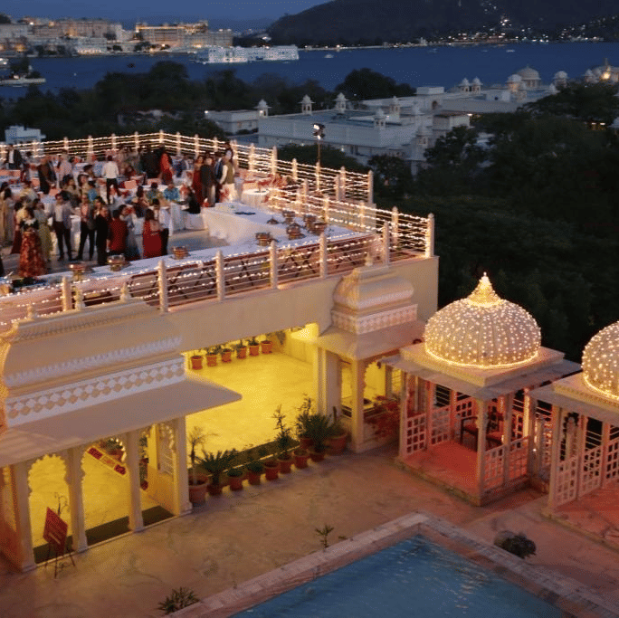 An event filled with guests on the upper terrace at Chunda Palace, Udaipur, overlooking the illuminated lower pool deck and the scenic Udaipur cityscape and lake in the distance.