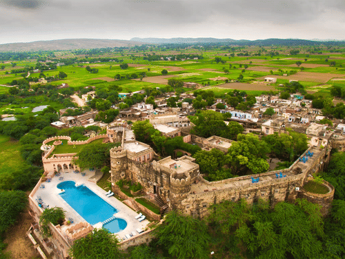 Aerial drone shot of Hill Fort Kesroli 14th Century Alwar where swimming pool and multiple structures are surrounded by lush green landscape and cloudy blue sky in the background.