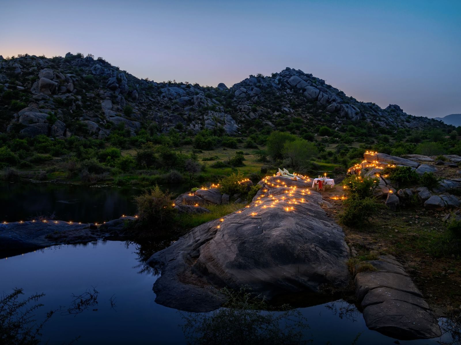 An image of an outdoor dining arrangement near a lake over viewing the mountains with bright lighting and greenery around - Utsav Camp Sariska