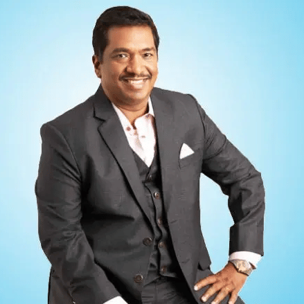 A man in a dark suit and tie, seated against a bright blue background - Accord Hotels & Resorts