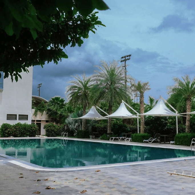 The swimming pool at the Arawali Ananta Elite Jaipur hotel with lounge chairs and umbrellas, surrounded by palm trees