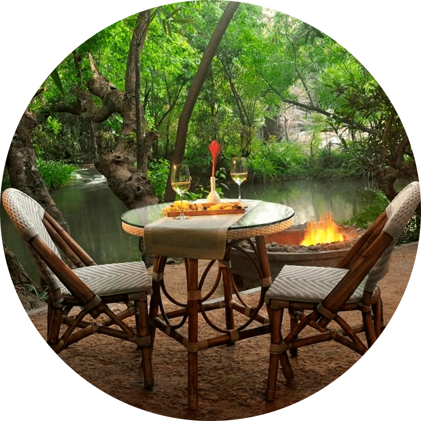 An outdoor dining setup with chairs for a couple, a small table, and a bonfire setup near a tranquil river in the forest at the Amanvana Spa Resort, Coorg - Luxury Resort
