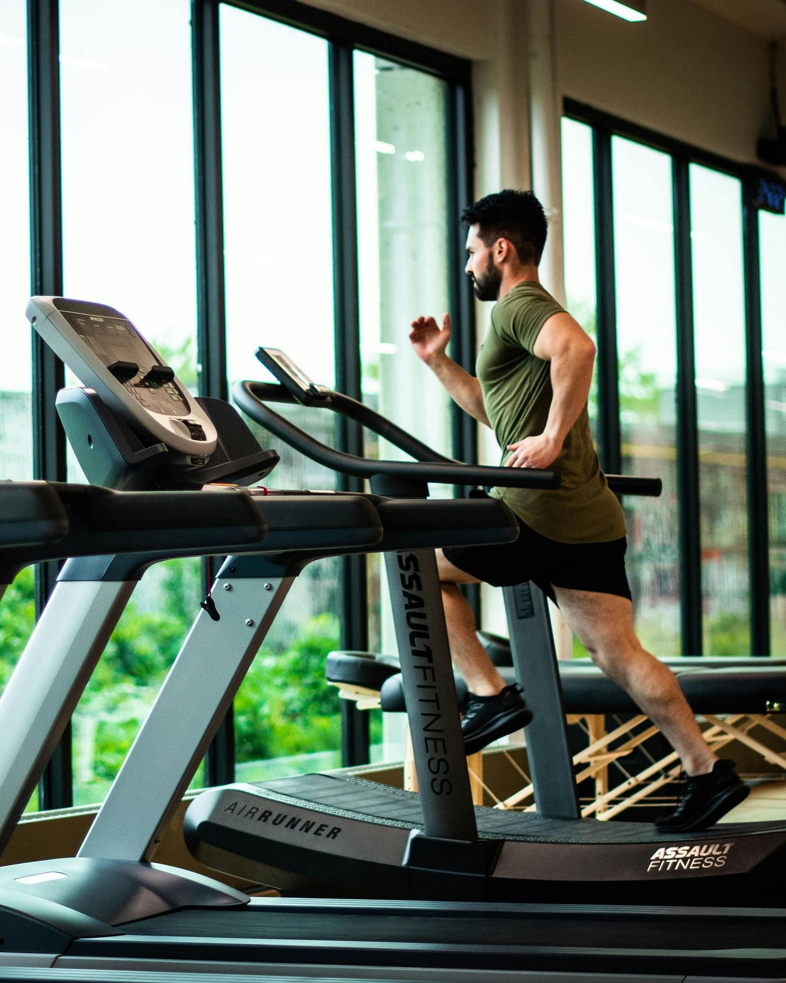 A person runs on a treadmill in a gym with large windows overlooking greenery. Other treadmills are in the foreground.