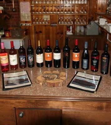 A collection of wine bottles is arranged on a wooden countertop, some with labels visible, along with two framed tasting notes and a wooden coaster, set against a wood-paneled background. - Kit Carson Lodge & Restaurant