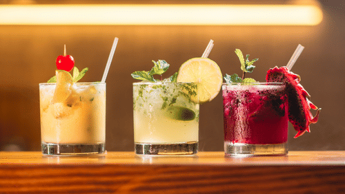 A row of four colourful cocktails garnished with fruit and herbs on a wooden bar.