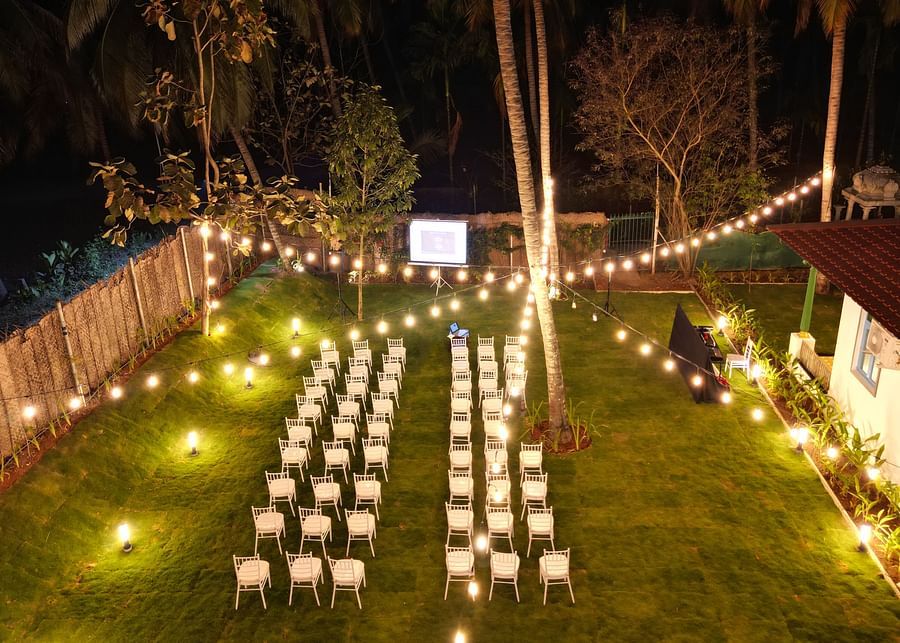 Outdoor night event setup with rows of white chairs, string lights, and a projector screen in a grassy yard.