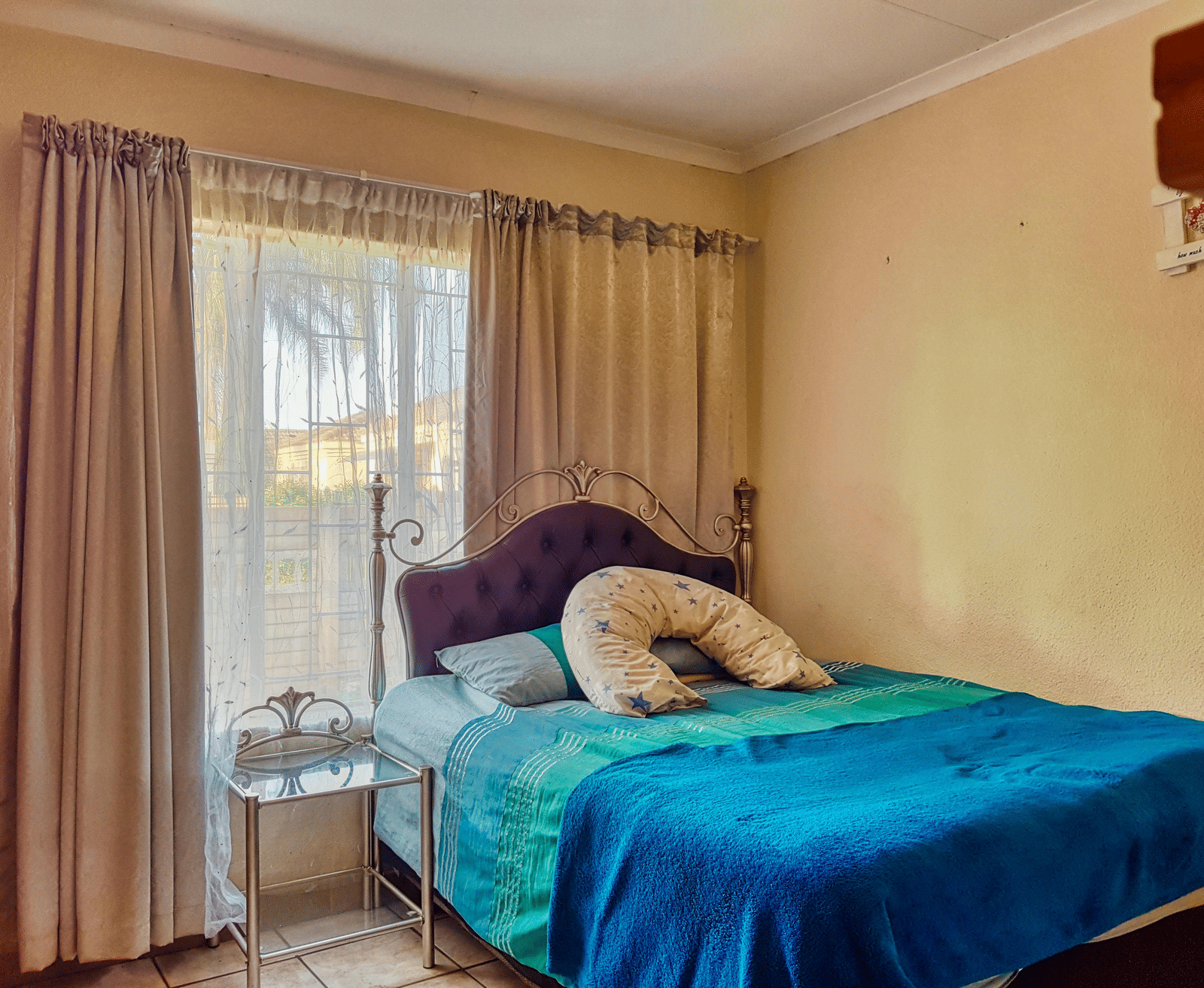 A bedroom featuring a double bed with blue sheets spread neatly over it with beige curtains covering the window behind it.