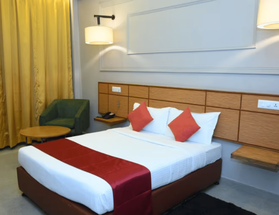 Bedroom with double bed, red runner, pillows, bedside lamps and yellow curtains at Max Hotel Prayagraj.