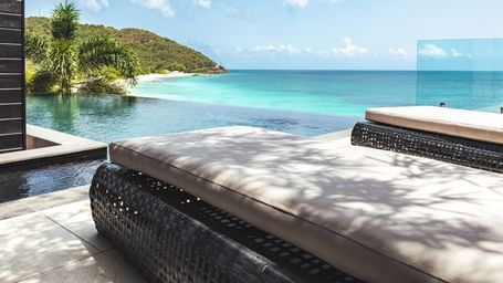 A tranquil infinity pool overlooks a pristine beach and turquoise ocean, with loungers in the foreground and green hills beyond - Tamarind Hills Resort and Villas - Antigua