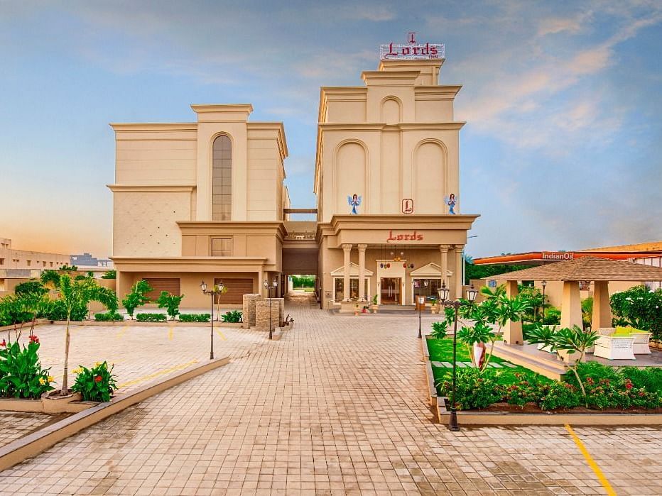 A wide shot of a light-coloured, multi-storey hotel building with a prominent entrance and surrounding greenery, including paved walkways and some parked cars, under a partly cloudy sky at either dawn or dusk at Lords Inn Motikhavdi, Jamnagar.