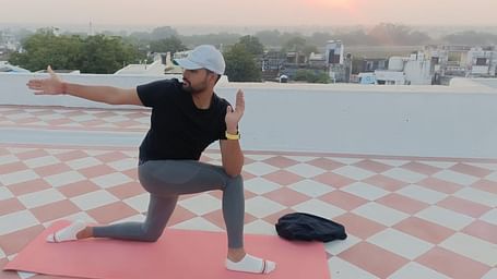 A person performs a yoga pose on a mat on a rooftop with a sunrise or sunset in the background.