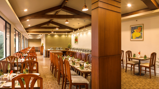 Restaurant interior showing a long row of tables and chairs. The room has large windows, wooden pillars, and exposed wooden beams - Abad Copper Castle, Munnar