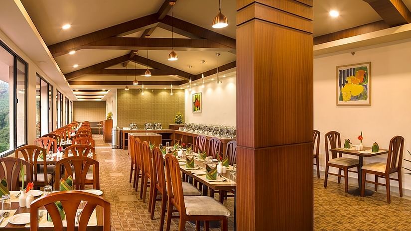 Restaurant interior showing a long row of tables and chairs. The room has large windows, wooden pillars, and exposed wooden beams - Abad Copper Castle, Munnar